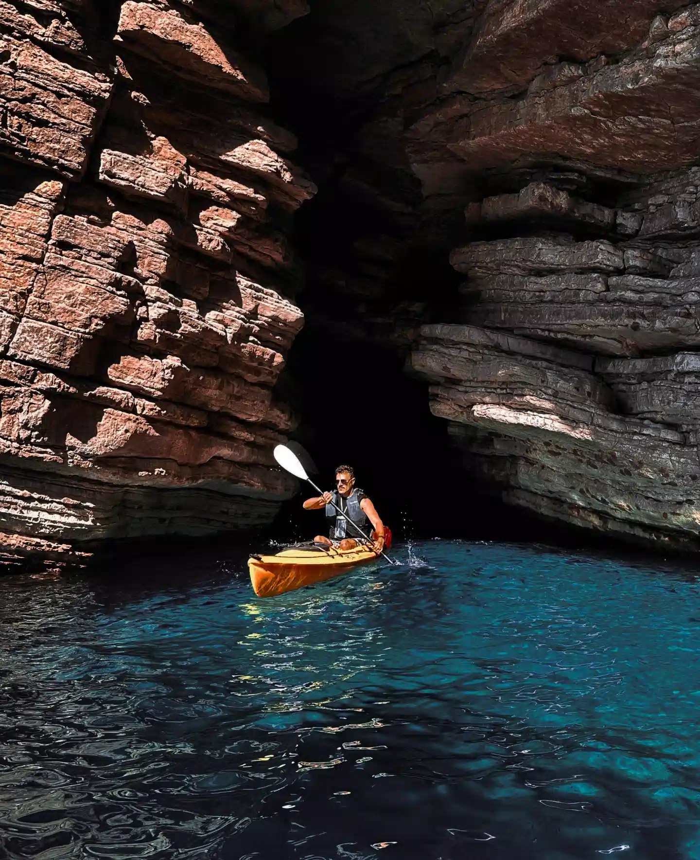 Man kayaking out of a rocky sea cave into bright blue water.