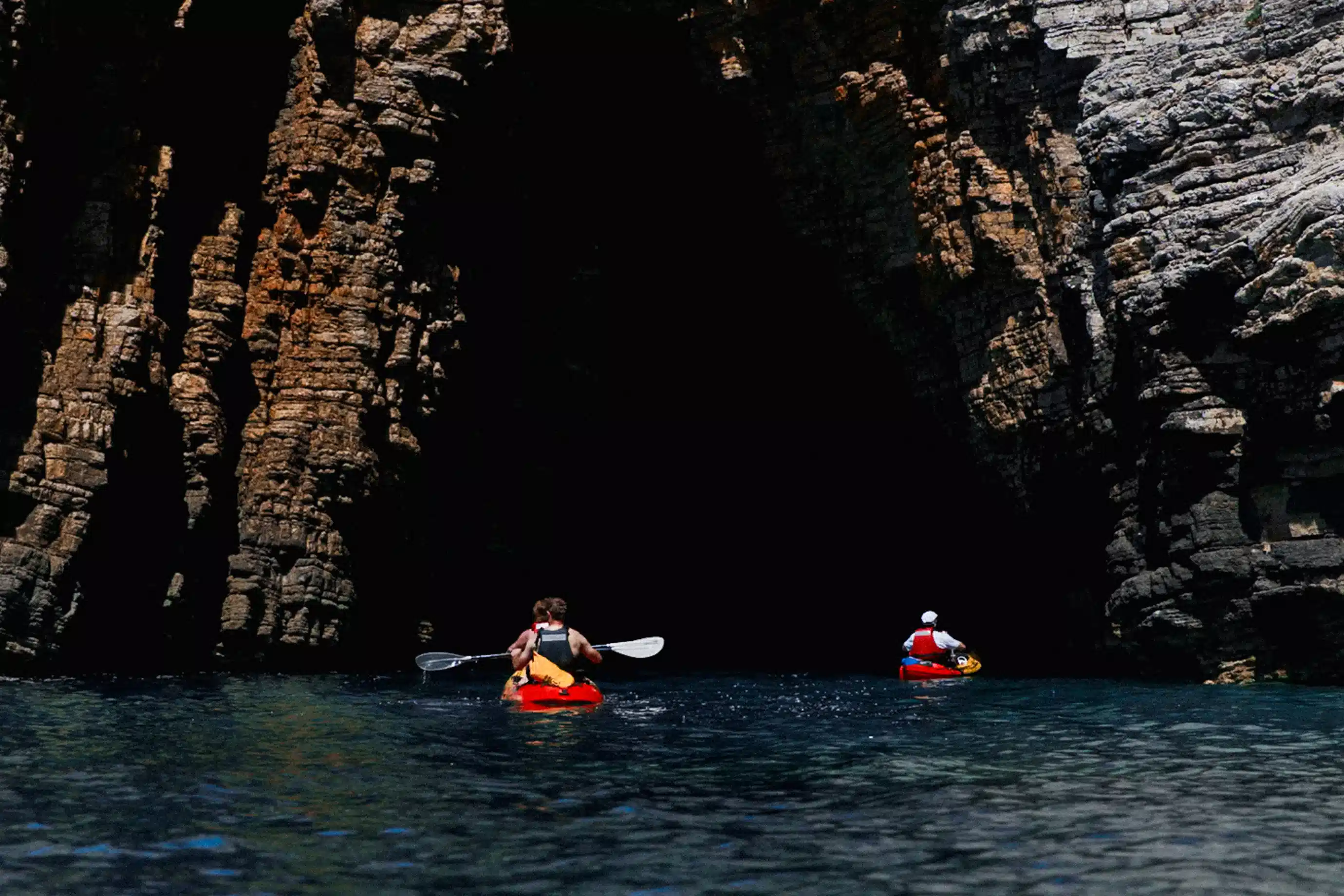 Two kayakers paddling toward the dark mouth of a coastal cave carved into towering limestone cliffs