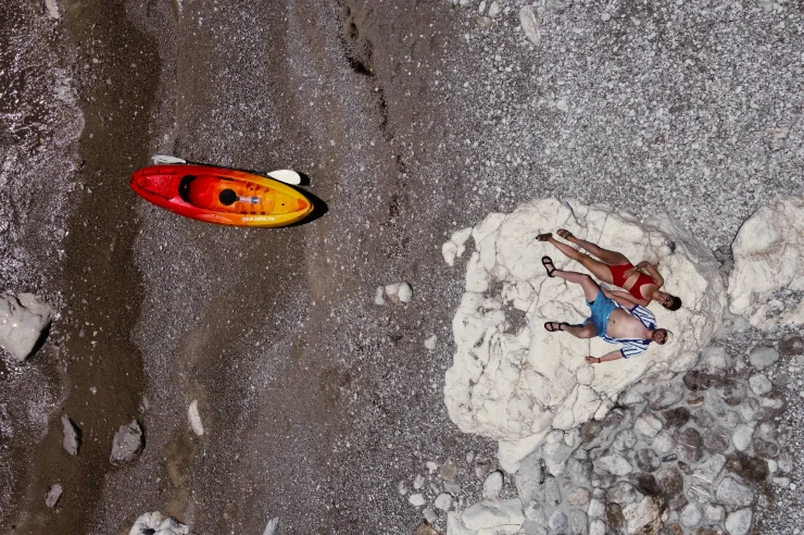 Aerial view of a red-and-yellow kayak pulled up on a pebble beach beside two sunbathers lying on a white limestone rock