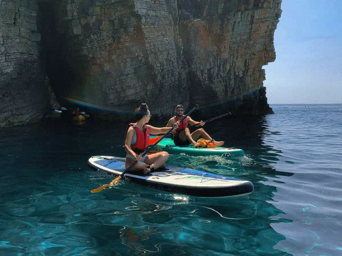 Two paddlers on SUP boards exploring a tall sea cliff with crystal-clear blue water