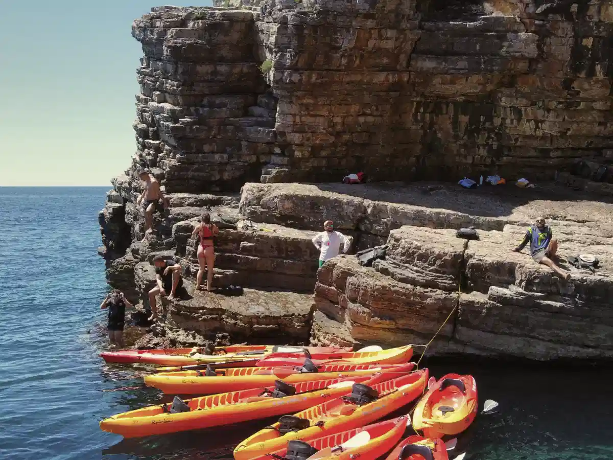 Group preparing seat-on-top kayaks at a rocky cliffside launch spot on a sunny day