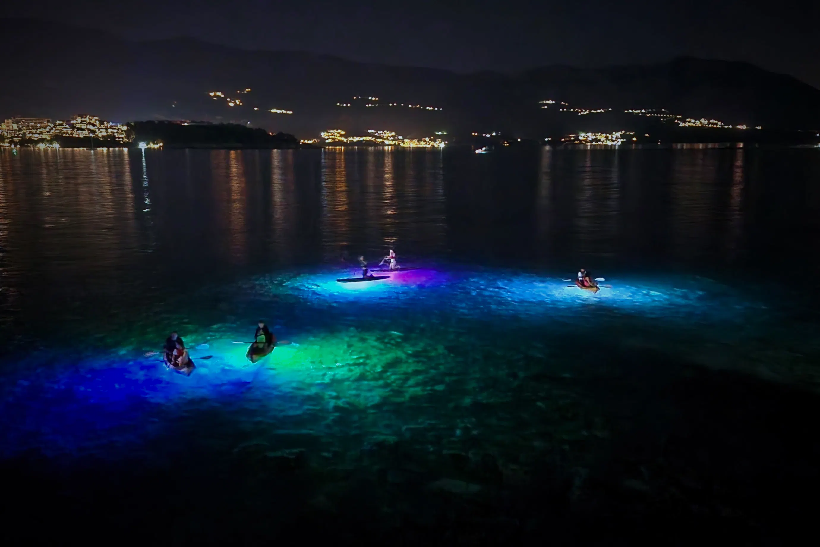 LED-lit kayaks floating over a bright blue glow with Budva skyline at night