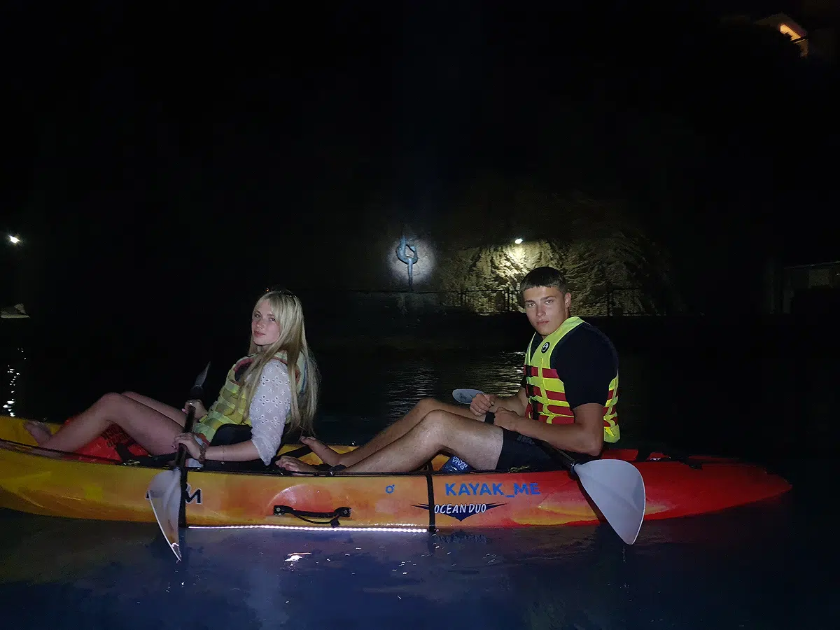 Couple paddling a double kayak by a rock wall at night, life jackets on – Night Light tour