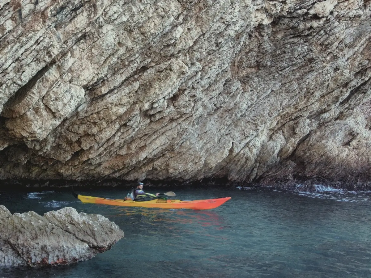 Person kayaking beneath a towering rock cliff in calm water