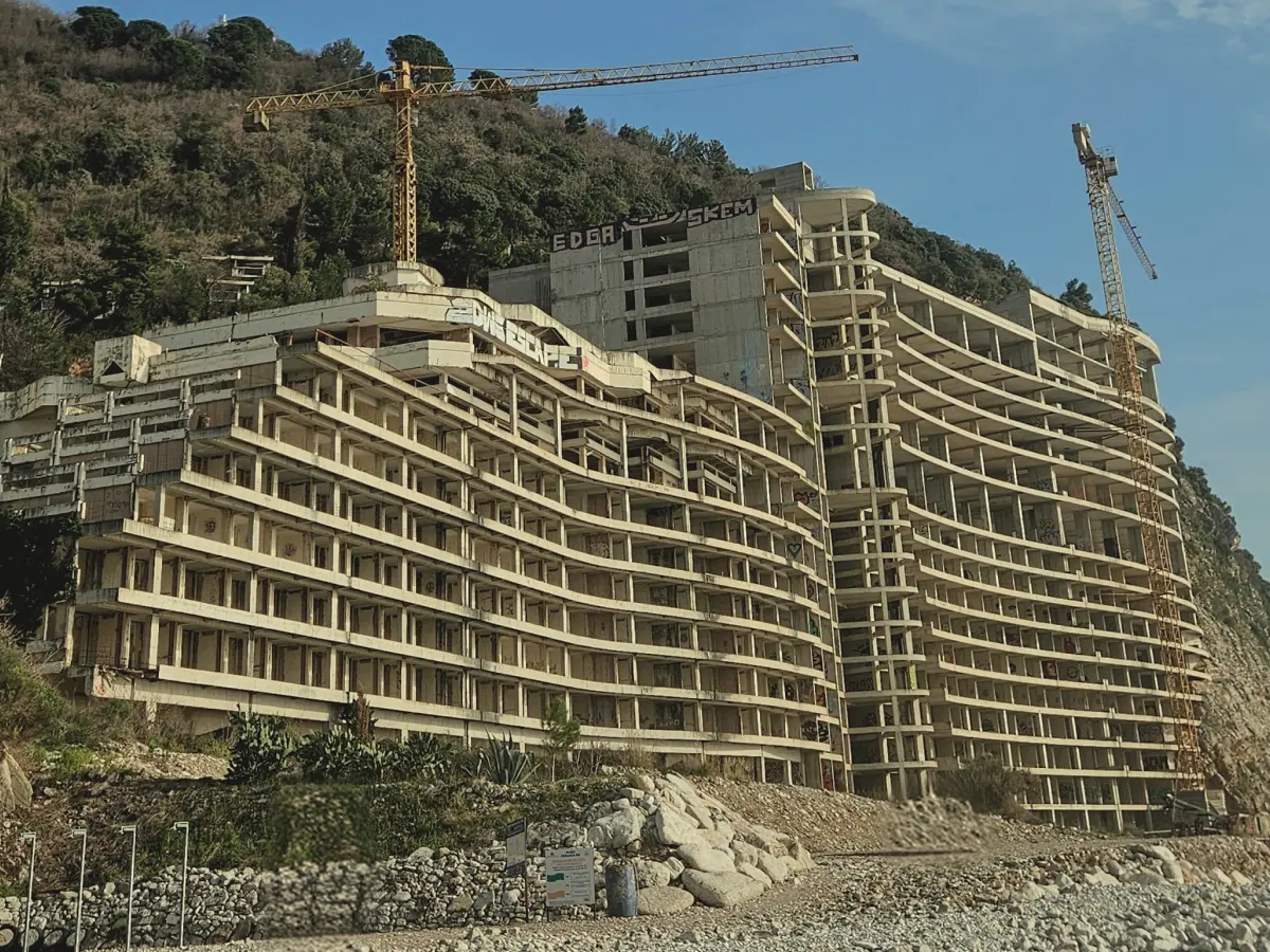 Large unfinished seaside building built into a steep hillside with cranes overhead